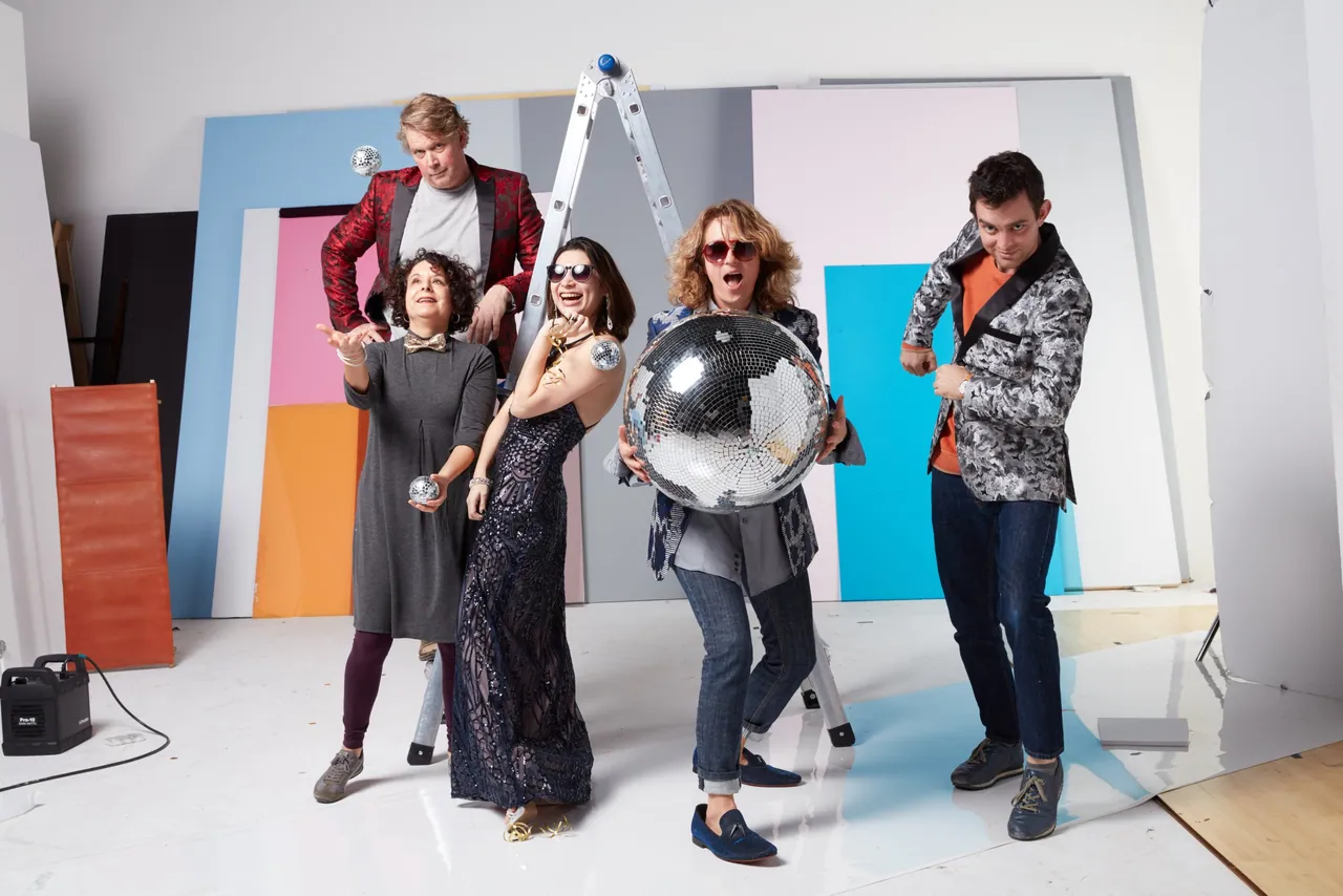 Team members posing with a giant disco ball and small mirror balls in front of colored studio panels