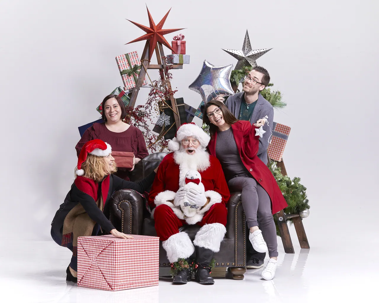 Team members posing around Santa in an armchair with gift boxes, stars and a wooden ladder tree