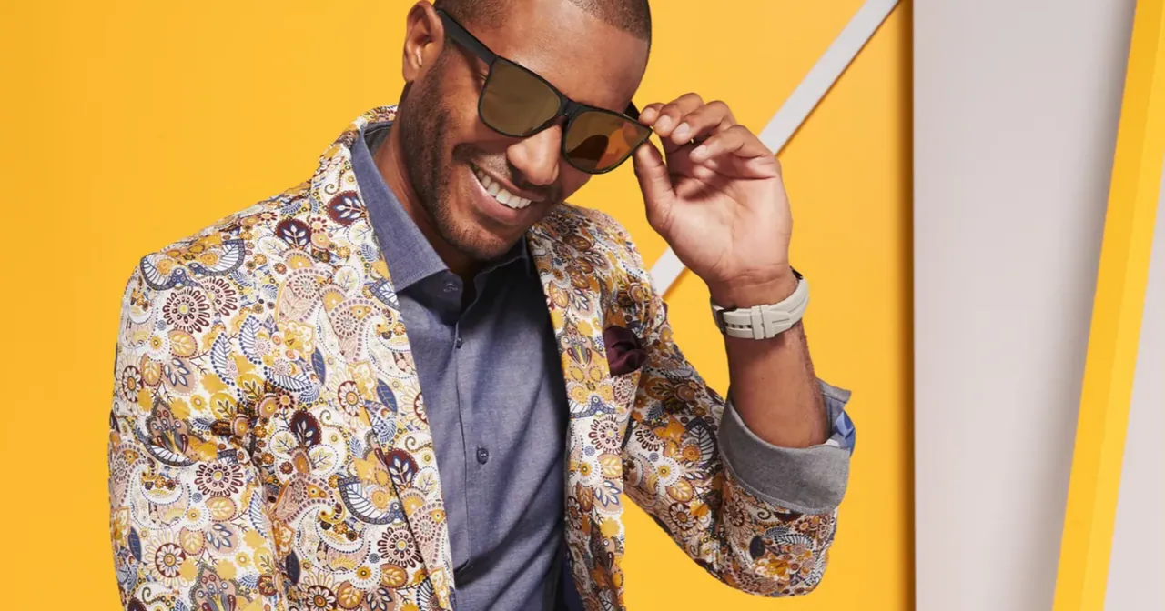 A smiling model in a paisley jacket and sunglasses on a yellow set