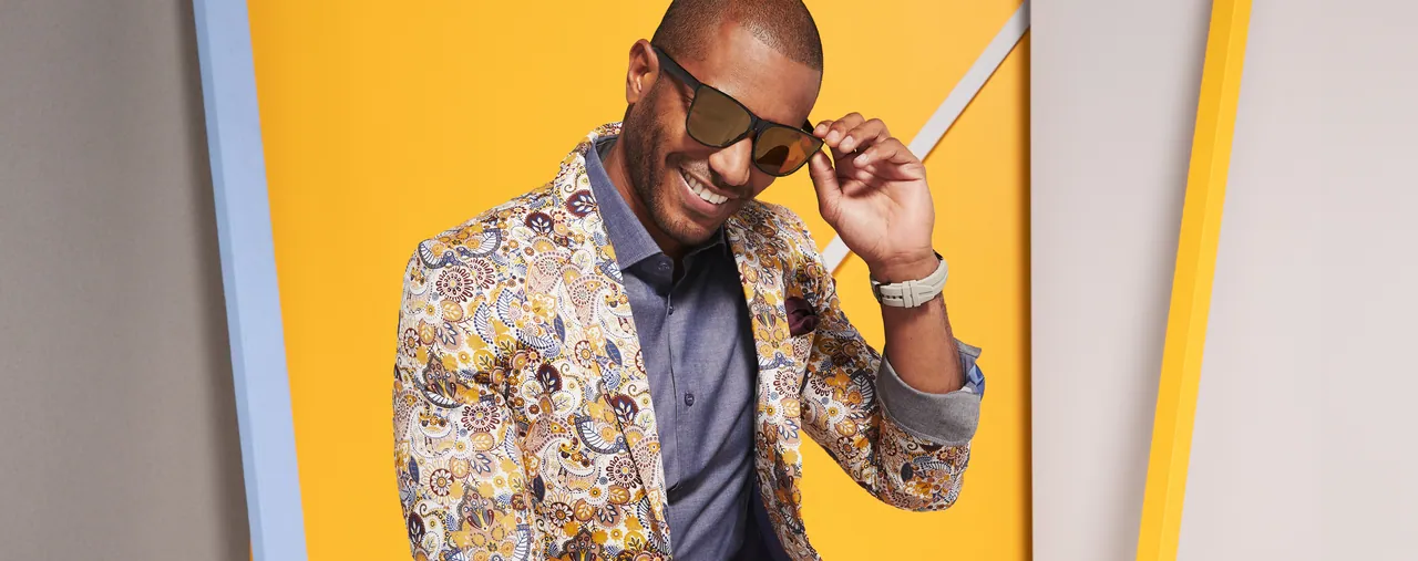 Model in a paisley jacket adjusting his sunglasses against a yellow set with a pale blue frame