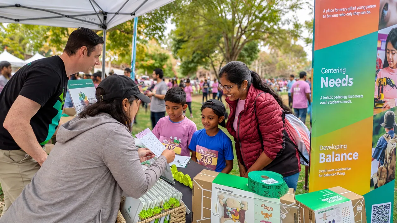 At a community event, two children and a parent look over admissions materials at the Tessellations table, beside the standing banner with the Centering Needs and Developing Balance bands