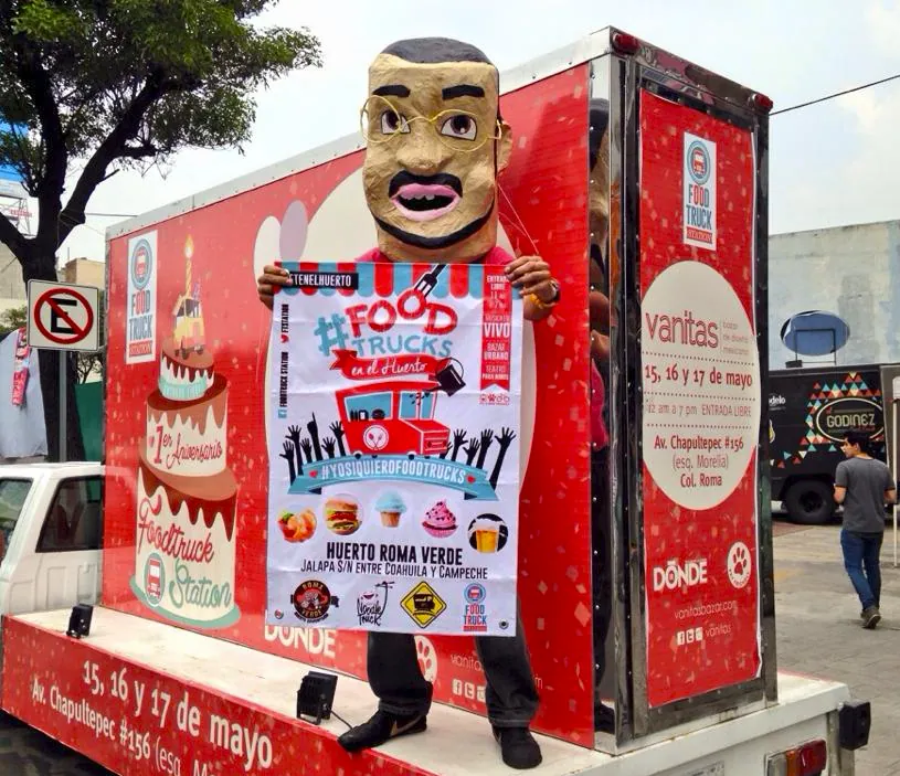 A costumed mascot holding an event poster in front of a red wrapped truck
